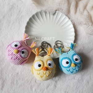 May include: Three crocheted owl keychains in pastel colors: pink, yellow, and blue. Each owl has large white eyes, a small orange beak, and decorative yarn tufts. The keychains are displayed on a white plate with the text "Nii CRAFT".