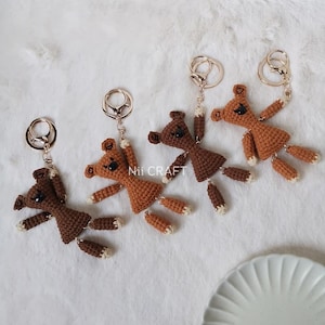 Può includere: Quattro portachiavi a forma di orsacchiotto di peluche in crochet marrone con anelli dorati. Gli orsacchiotti hanno occhi neri e naso marrone. I portachiavi sono disposti in fila su una superficie bianca. Il testo "Nii CRAFT" è visibile nell'immagine.