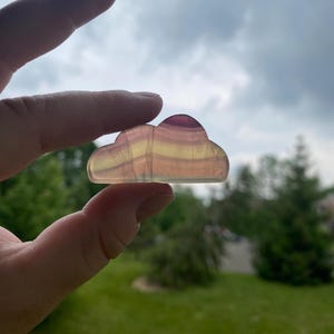 May include: A translucent, cloud-shaped crystal with layers of purple, yellow, and pink hues. The polished stone is held up against a backdrop of a cloudy sky and green foliage.