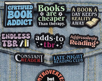 May include: Nine enamel pins with book-themed sayings. The pins are black with colorful text and images. The sayings include "Certified Book Addict", "Books are cheaper than therapy", "A book a day keeps reality away", "Endless TBR", "Adds to TBR", "Aggressively Reading", "Constant Reader", "Late Night Reading Club", and "Introverted but willing to discuss books".