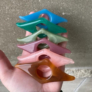 May include: A collection of colorful, acrylic thumb page holders in shades of blue, green, pink, and brown. Each holder has a diamond shape with a circular opening for the thumb, designed to keep pages open while reading.