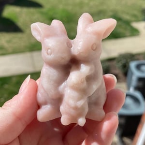 May include: A light pink stone carving of three rabbits. The two larger rabbits are embracing, with a smaller rabbit nestled between them. The carving is held in a hand, with a blurred outdoor background.
