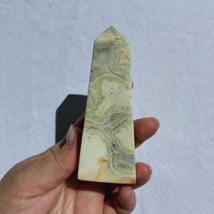 Puede incluir: Un obelisco de cristal rectangular y alto, con una parte superior puntiaguda. La piedra presenta una base de color crema con patrones arremolinados verdes y grises. El obelisco se sostiene en una mano sobre un fondo blanco.