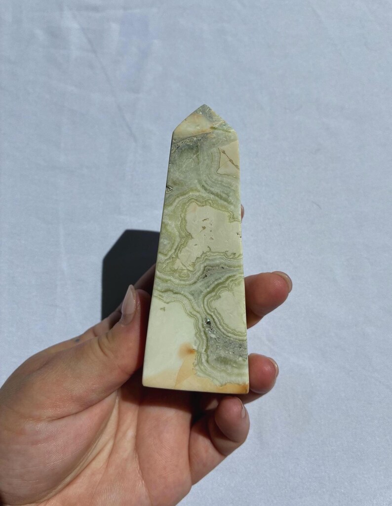 Puede incluir: Un obelisco de cristal rectangular y alto, con una parte superior puntiaguda. La piedra presenta una base de color crema con patrones arremolinados verdes y grises. El obelisco se sostiene en una mano sobre un fondo blanco.