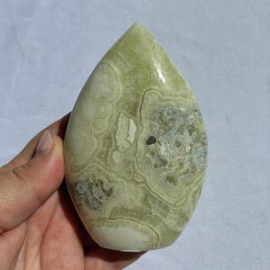 Puede incluir: Una piedra pulida en forma de l&aacute;grima, con un tono verde claro y patrones arremolinados intrincados. La piedra tiene &aacute;reas blancas y verde oscuro, con un acabado liso y brillante. Un art&iacute;culo decorativo natural.