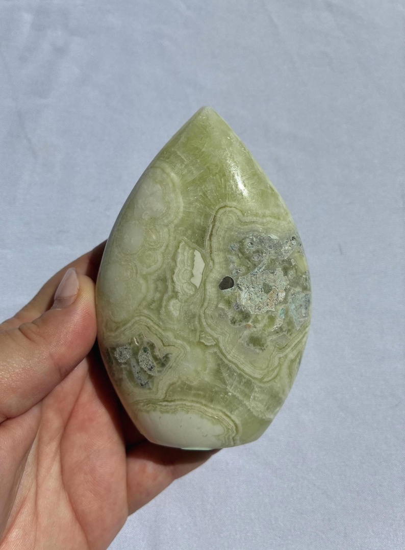 Puede incluir: Una piedra pulida en forma de l&aacute;grima, con un tono verde claro y patrones arremolinados intrincados. La piedra tiene &aacute;reas blancas y verde oscuro, con un acabado liso y brillante. Un art&iacute;culo decorativo natural.
