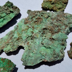May include: Close-up of several pieces of raw malachite mineral. The malachite has a vibrant green color with areas of brown and a rough, textured surface. The pieces vary in size and shape, with some appearing more crystalline.