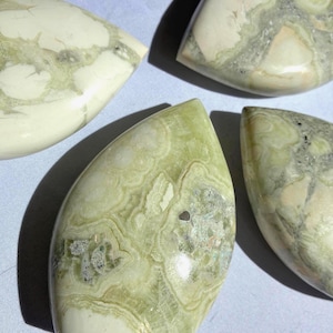 Puede incluir: Cuatro piedras pulidas en forma de l&aacute;grima con apariencia veteada. Las piedras presentan una mezcla de tonos verde p&aacute;lido, blanco y gris, con intrincados patrones arremolinados. Las superficies son lisas y brillantes, reflejando la luz.