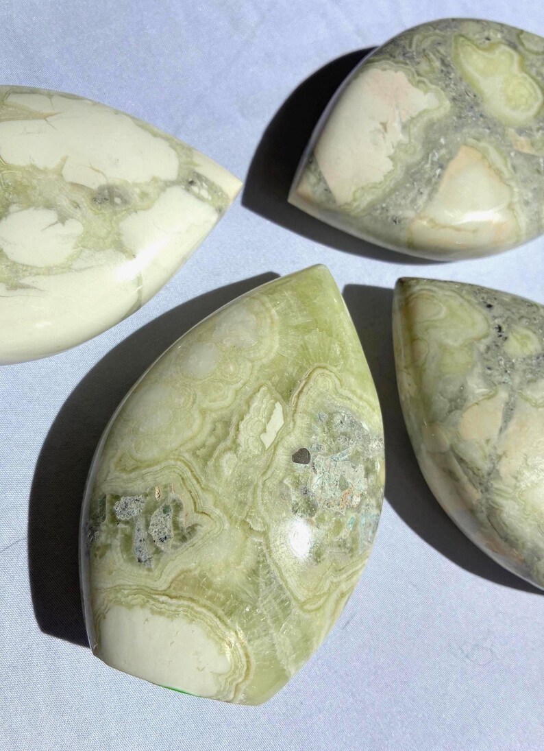 Puede incluir: Cuatro piedras pulidas en forma de l&aacute;grima con apariencia veteada. Las piedras presentan una mezcla de tonos verde p&aacute;lido, blanco y gris, con intrincados patrones arremolinados. Las superficies son lisas y brillantes, reflejando la luz.