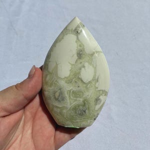 Puede incluir: Una piedra en forma de l&aacute;grima con una superficie pulida. La piedra presenta un color base blanco crema con intrincados patrones de vetas verdes y grises. La piedra se sostiene en una mano.