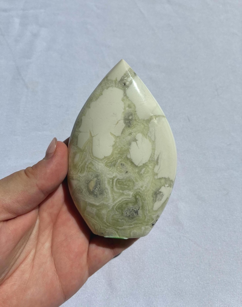 Puede incluir: Una piedra en forma de l&aacute;grima con una superficie pulida. La piedra presenta un color base blanco crema con intrincados patrones de vetas verdes y grises. La piedra se sostiene en una mano.