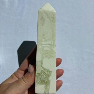 Puede incluir: Un obelisco de cristal rectangular con una parte superior puntiaguda. La piedra es blanquecina con vetas y patrones verdes y verde claro. El obelisco se sostiene en una mano sobre un fondo blanco.