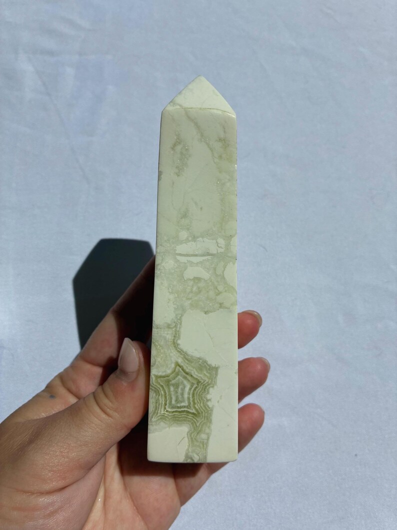 Puede incluir: Un obelisco de cristal rectangular con una parte superior puntiaguda. La piedra es blanquecina con vetas y patrones verdes y verde claro. El obelisco se sostiene en una mano sobre un fondo blanco.