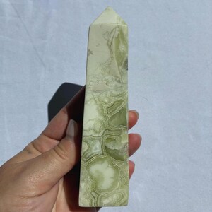 Puede incluir: Un obelisco de piedra pulida, alto y rectangular. La piedra tiene un patr&oacute;n veteado en verde claro y blanco. La parte superior del obelisco tiene forma de pir&aacute;mide puntiaguda. El obelisco est&aacute; sostenido en una mano.