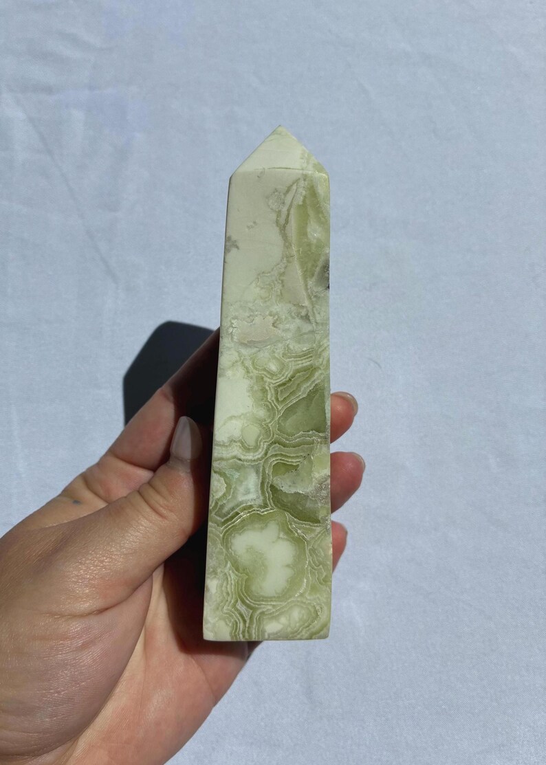Puede incluir: Un obelisco de piedra pulida, alto y rectangular. La piedra tiene un patr&oacute;n veteado en verde claro y blanco. La parte superior del obelisco tiene forma de pir&aacute;mide puntiaguda. El obelisco est&aacute; sostenido en una mano.