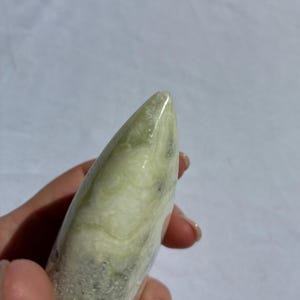 Puede incluir: Una piedra pulida de color verde claro, sostenida en una mano. La piedra tiene una forma lisa y redondeada con una punta. El color es verde p&aacute;lido con vetas verdes y grises m&aacute;s oscuras.