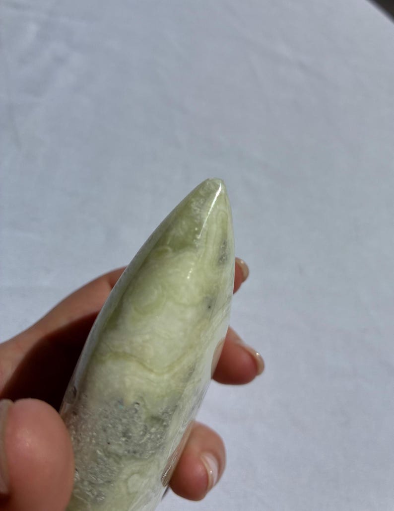 Puede incluir: Una piedra pulida de color verde claro, sostenida en una mano. La piedra tiene una forma lisa y redondeada con una punta. El color es verde p&aacute;lido con vetas verdes y grises m&aacute;s oscuras.