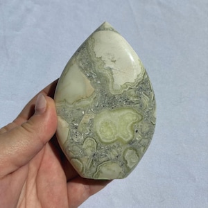Puede incluir: Una piedra pulida en forma de l&aacute;grima con una mezcla de patrones verde p&aacute;lido, crema y gris. La piedra tiene una superficie lisa y brillante y una parte superior puntiaguda. La apariencia general es org&aacute;nica y natural, con un dise&ntilde;o veteado &uacute;nico.