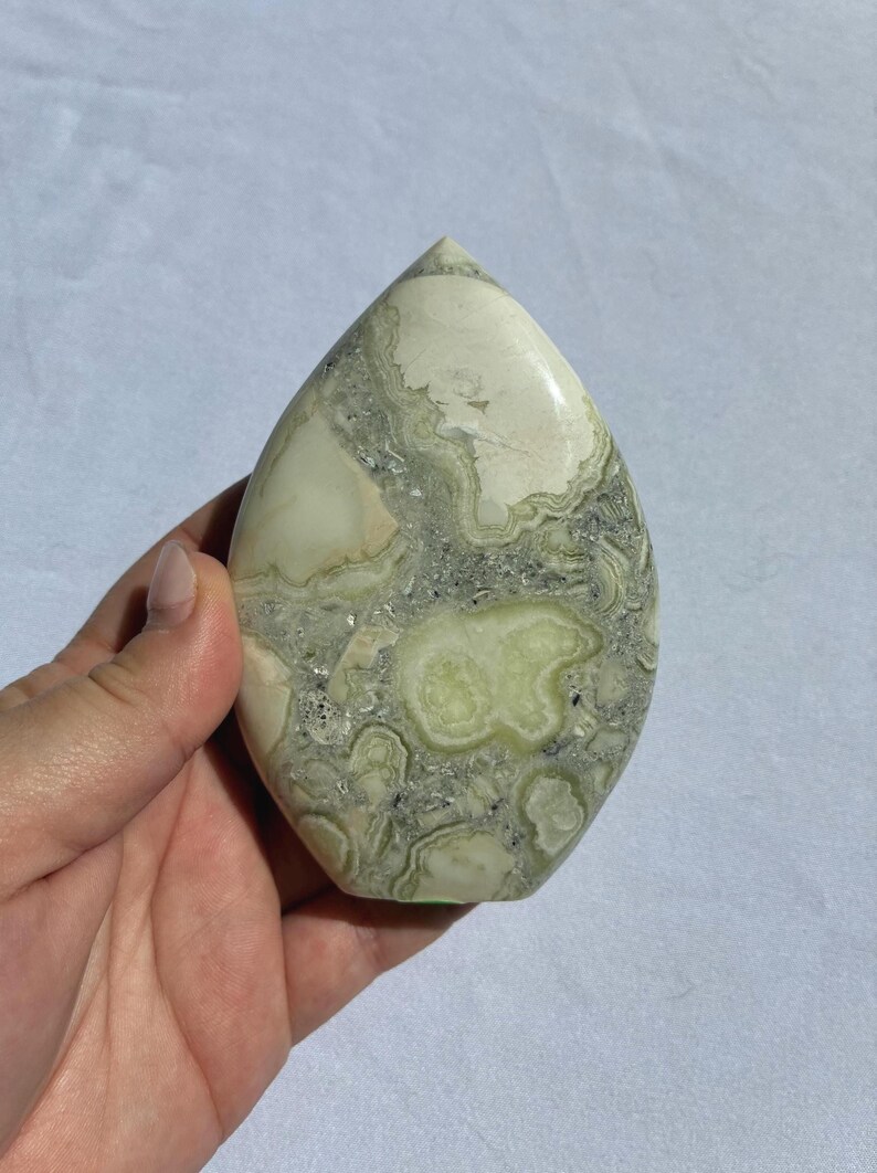 Puede incluir: Una piedra pulida en forma de l&aacute;grima con una mezcla de patrones verde p&aacute;lido, crema y gris. La piedra tiene una superficie lisa y brillante y una parte superior puntiaguda. La apariencia general es org&aacute;nica y natural, con un dise&ntilde;o veteado &uacute;nico.