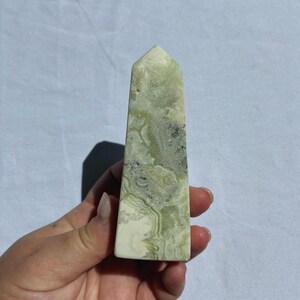 Puede incluir: Un obelisco de cristal rectangular pulido. La piedra presenta una mezcla de patrones verde p&aacute;lido, blanco y gris. El obelisco se sostiene en una mano sobre un fondo blanco. La parte superior del obelisco tiene forma de pir&aacute;mide puntiaguda.