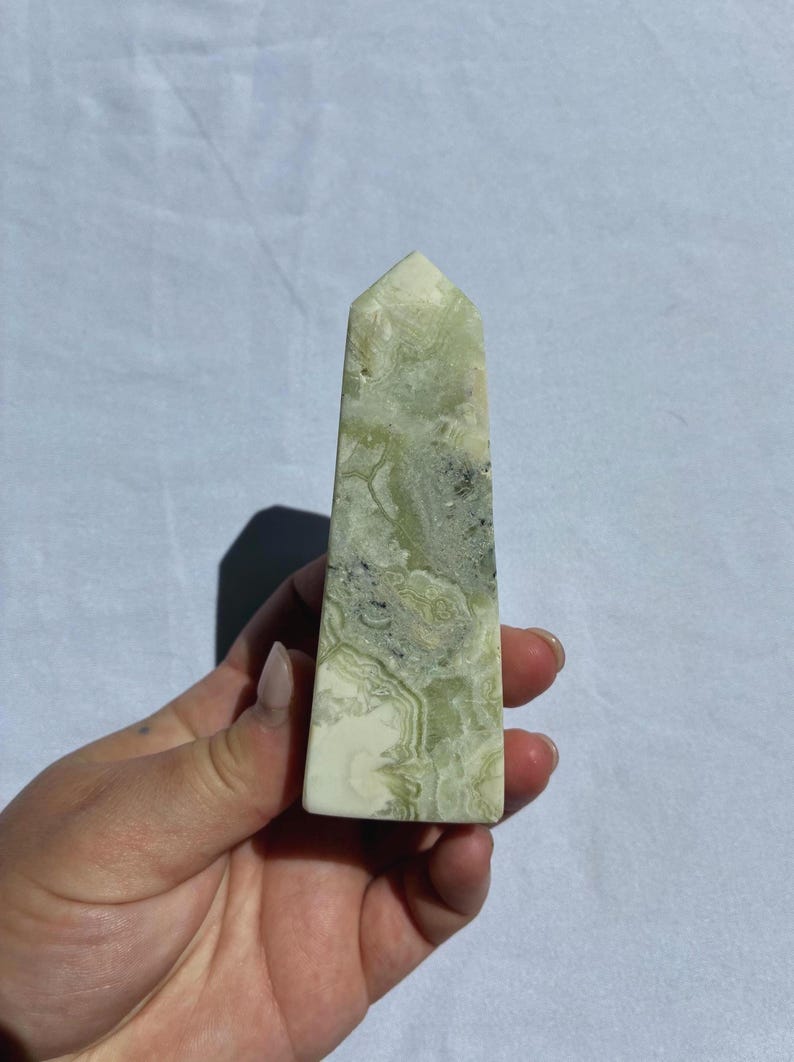 Puede incluir: Un obelisco de cristal rectangular pulido. La piedra presenta una mezcla de patrones verde p&aacute;lido, blanco y gris. El obelisco se sostiene en una mano sobre un fondo blanco. La parte superior del obelisco tiene forma de pir&aacute;mide puntiaguda.