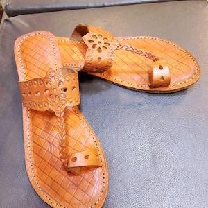May include: A pair of brown leather sandals with intricate cut-out designs and braided straps. The sandals have a square-shaped toe and a flat sole.