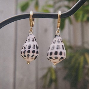 Puede incluir: Un par de pendientes colgantes con pequeñas conchas en forma de pera, con una base blanca y patrones de manchas rectangulares negras. Cada concha está suspendida de un cierre de aro de metal en tono dorado, creando un diseño de joyería sencillo.