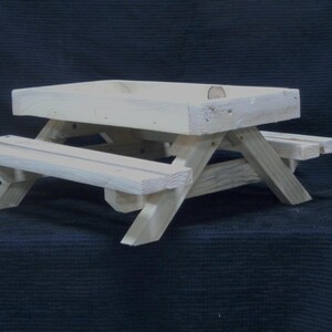 May include: A small, unfinished wooden picnic table with a rectangular top and attached benches. The table is constructed from light-colored wood and features a classic picnic table design. Ideal for small toys or decorative items.