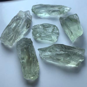 Puede incluir: Seis cristales translúcidos de color verde pálido de forma irregular están dispersos sobre una superficie clara. Los cristales tienen bordes ásperos y dentados y tamaños variables, capturando la luz para crear reflejos y sombras. La apariencia general sugiere piedras preciosas naturales en bruto.