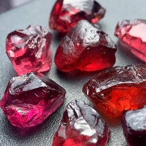 Puede incluir: Un conjunto de piedras preciosas de granate en bruto, sin cortar. Las gemas varían en tamaño y forma, mostrando un tono rojo intenso con diferentes grados de transparencia. Se aprecian la textura rugosa y las formaciones naturales de las piedras.