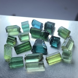 Puede incluir: Una colección de cristales de turmalina en bruto en varios tonos de verde y azul. Las piedras son alargadas, prismáticas y presentan estrías naturales a lo largo de su longitud, mostrando una mezcla de degradados de color translúcidos y opacos.
