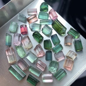 May include: A collection of small, raw, multi-colored tourmaline crystals scattered on a silver-toned metal tray. The stones feature various shades of green, pink, blue, and clear hues, displaying natural vertical striations and prismatic shapes.