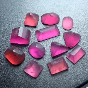 Può includere: Una collezione di gemme sfaccettate in tonalità rosa e rosse. Le gemme variano nella forma, inclusi tagli rettangolari, quadrati e ovali. Le pietre sono traslucide, con un aspetto lucido, e sono disposte su una superficie scura.