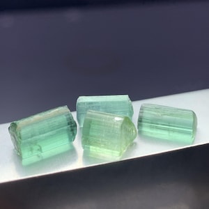 May include: Four small, translucent, light green tourmaline crystals with a prismatic, elongated shape and vertical striations along their sides. The crystals exhibit a clear, glassy texture and are arranged in a cluster, showing natural facets.