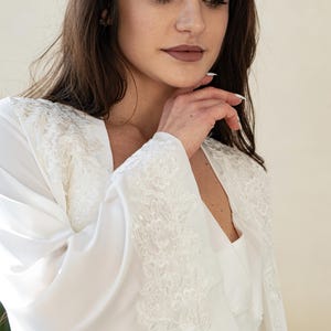 May include: A white satin robe with intricate lace detailing on the sleeves and shoulders. The robe has a V-neck design and is made of a soft, flowing fabric. The image shows a close-up of the robe, highlighting the delicate lace and the smooth texture of the satin.