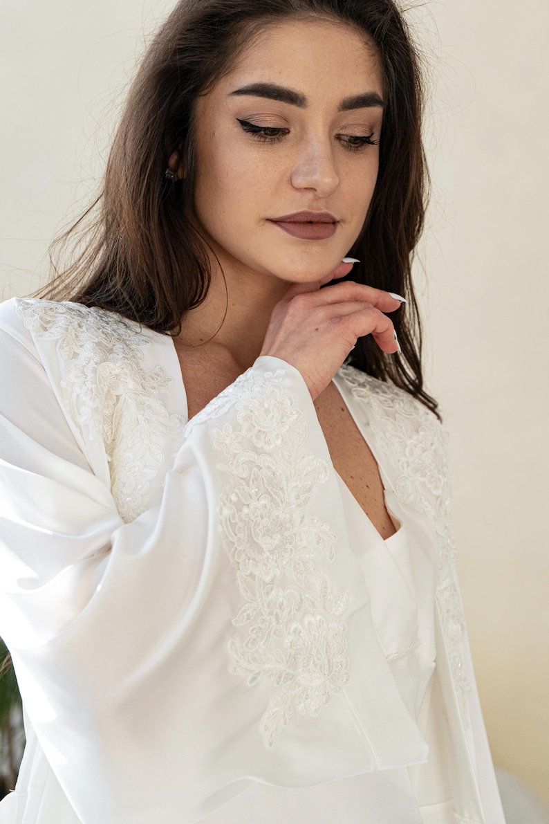 May include: A white satin robe with intricate lace detailing on the sleeves and shoulders. The robe has a V-neck design and is made of a soft, flowing fabric. The image shows a close-up of the robe, highlighting the delicate lace and the smooth texture of the satin.