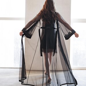 May include: A sheer black robe with a dotted pattern and lace trim. The robe has long sleeves and a matching belt. The model is wearing a black slip dress and silver heels.