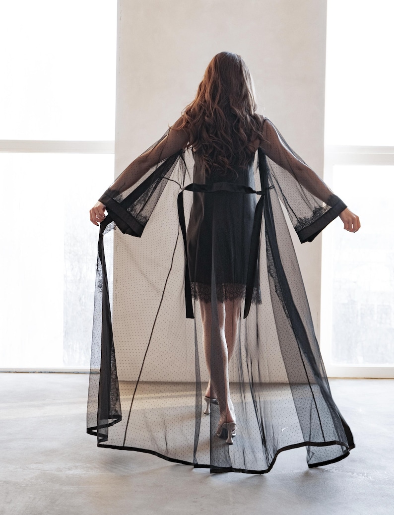 May include: A sheer black robe with a dotted pattern and lace trim. The robe has long sleeves and a matching belt. The model is wearing a black slip dress and silver heels.