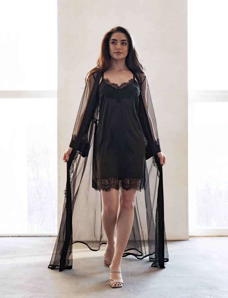 May include: A black lingerie set featuring a slip dress with lace trim and a sheer, dotted robe. The robe has long sleeves and lace detailing. The set is designed for sleepwear or loungewear.
