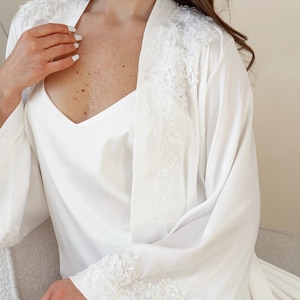 May include: A white satin robe and slip set. The robe features delicate lace detailing along the sleeves and shoulders. The V-neck slip is visible beneath the robe. The set is designed for comfort and elegance.