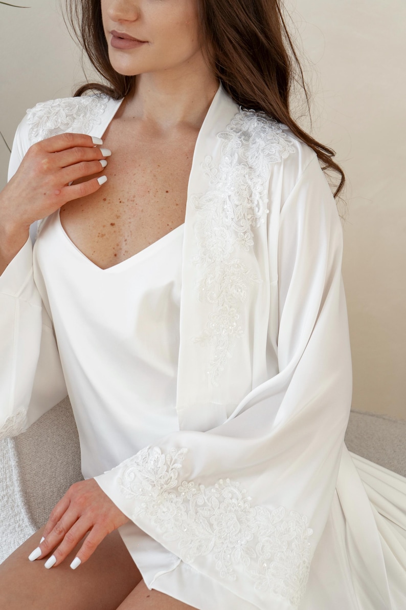 May include: A white satin robe and slip set. The robe features delicate lace detailing along the sleeves and shoulders. The V-neck slip is visible beneath the robe. The set is designed for comfort and elegance.