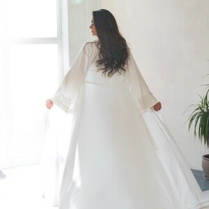 May include: A long, flowing white robe with wide sleeves and delicate lace detailing on the cuffs. The robe is worn by a person with long, dark hair, standing near a window. The overall aesthetic is soft and elegant, suggesting bridal or loungewear.