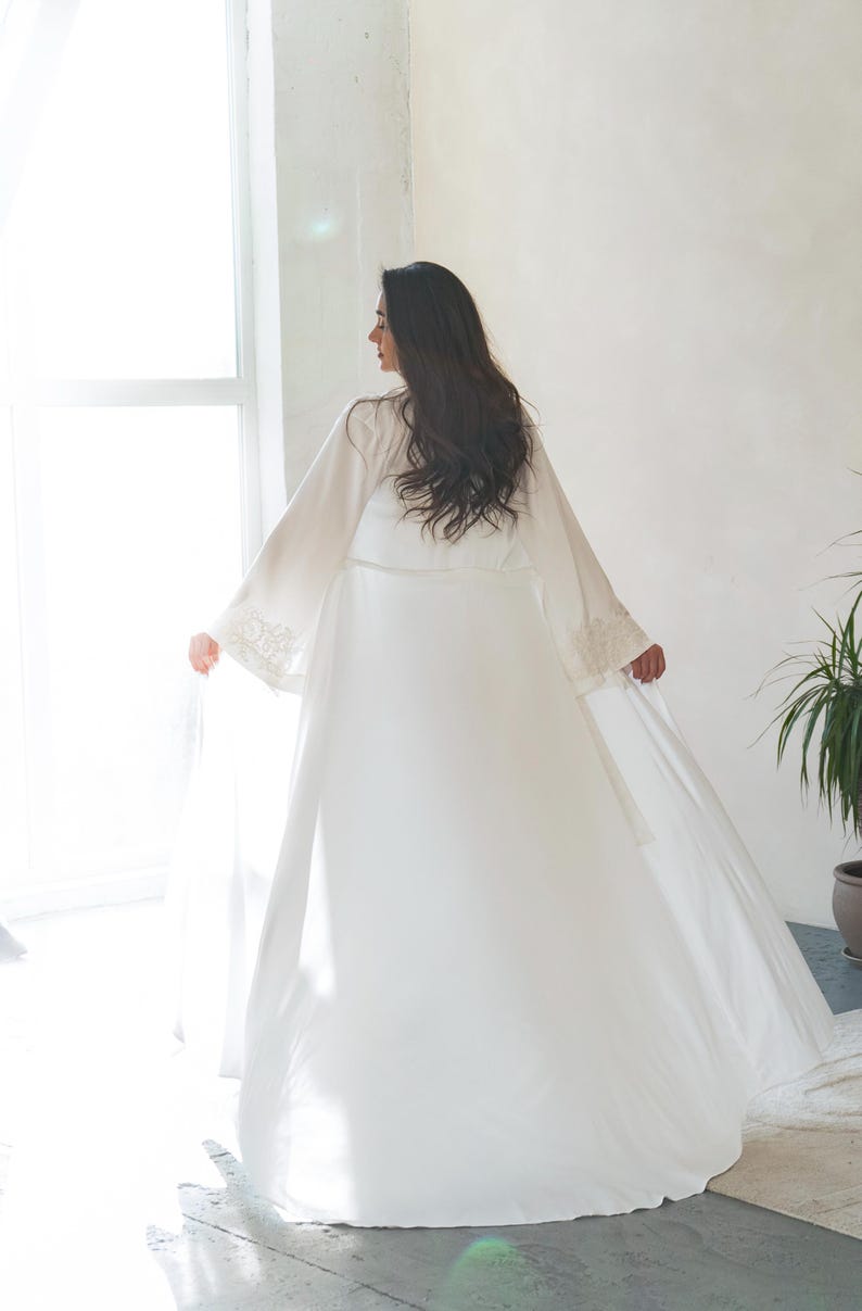 May include: A long, flowing white robe with wide sleeves and delicate lace detailing on the cuffs. The robe is worn by a person with long, dark hair, standing near a window. The overall aesthetic is soft and elegant, suggesting bridal or loungewear.