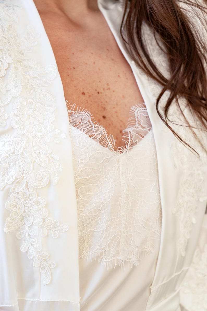 May include: White satin robe with delicate lace detailing. The robe features a V-neckline and long sleeves.