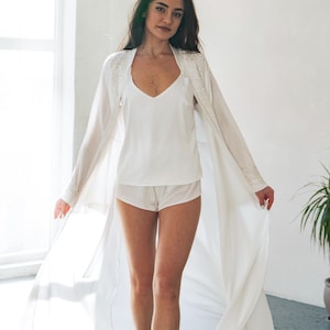 May include: A white lingerie set featuring a camisole, shorts, and a long, flowing robe. The robe has lace detailing on the shoulders and sleeves. The outfit is made of a soft, lightweight fabric.