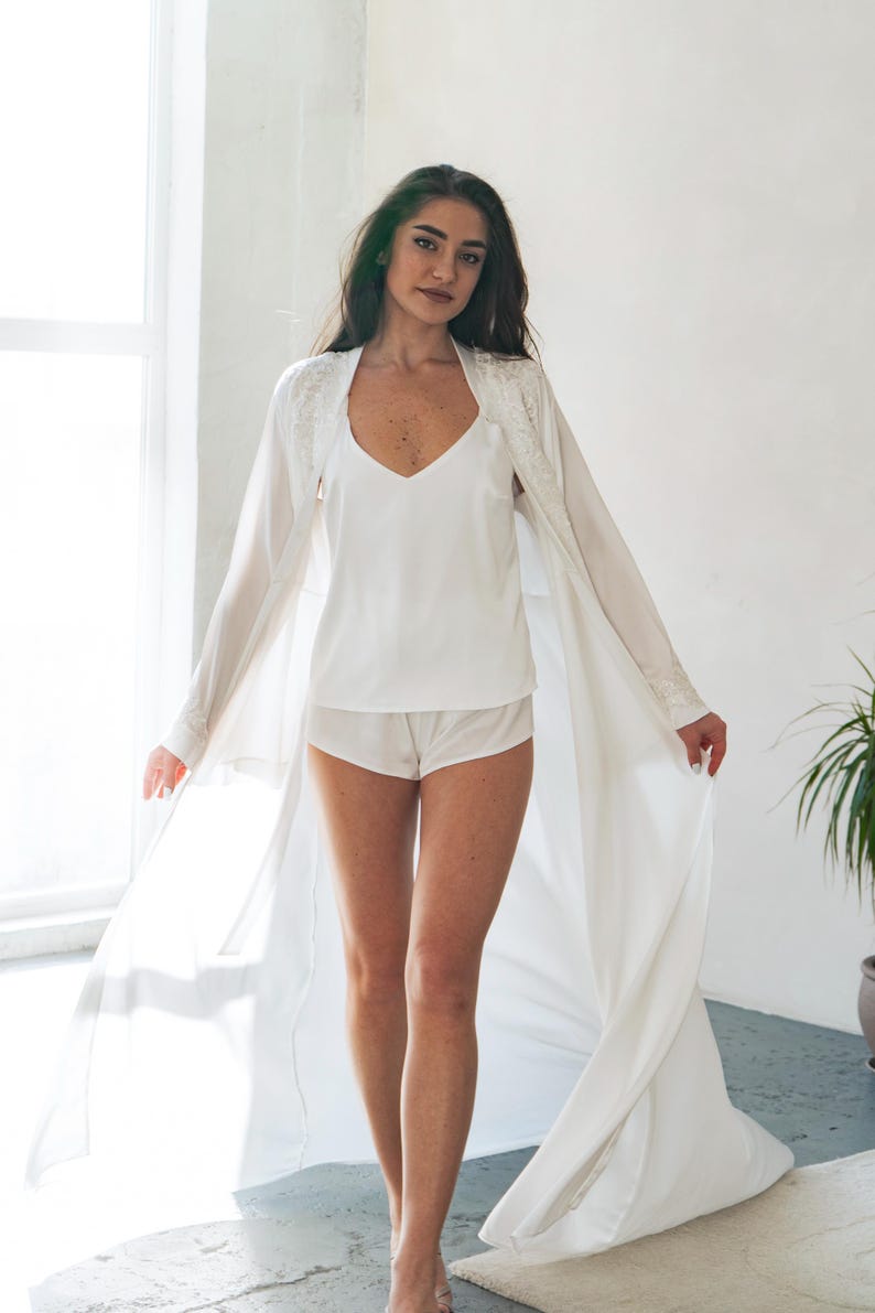 May include: A white lingerie set featuring a camisole, shorts, and a long, flowing robe. The robe has lace detailing on the shoulders and sleeves. The outfit is made of a soft, lightweight fabric.