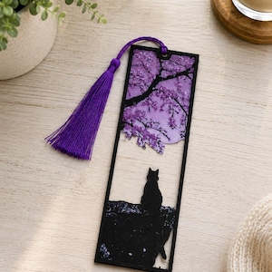Puede incluir: Marcapáginas decorativo con una silueta de gato negro sobre un fondo floral púrpura. El marcapáginas tiene un borde negro y una borla púrpura. El diseño incluye una rama de árbol y una luna llena.