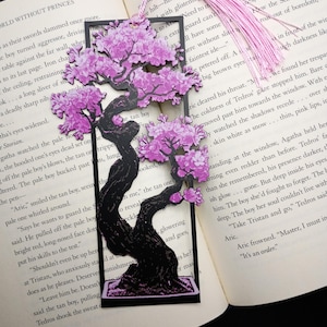 Puede incluir: Marcapáginas negro con un diseño de bonsái y flores de cerezo rosas, con una borla rosa. El marcapáginas está en un libro abierto.