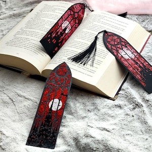 Puede incluir: Marcadores de estilo gótico con un diseño de vidriera roja y negra que presenta una luna llena y murciélagos. Los marcadores se muestran con un libro abierto sobre una superficie suave y texturizada. Cada marcador tiene una borla negra.