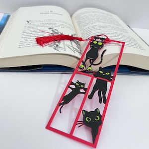 Op de afbeelding: Een rood omlijnde bladwijzer met zwarte katten silhouetten en groene ogen, rustend op een open boek. De bladwijzer heeft vier kattenontwerpen in verschillende poses. Een rode kwast hangt aan de bovenkant van het boek.